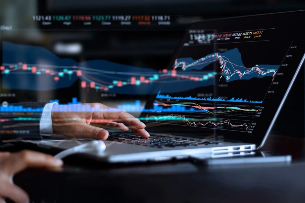 A person analyzes financial data on a laptop showing colorful stock market charts, focused on keyboard input with a smartphone beside them.