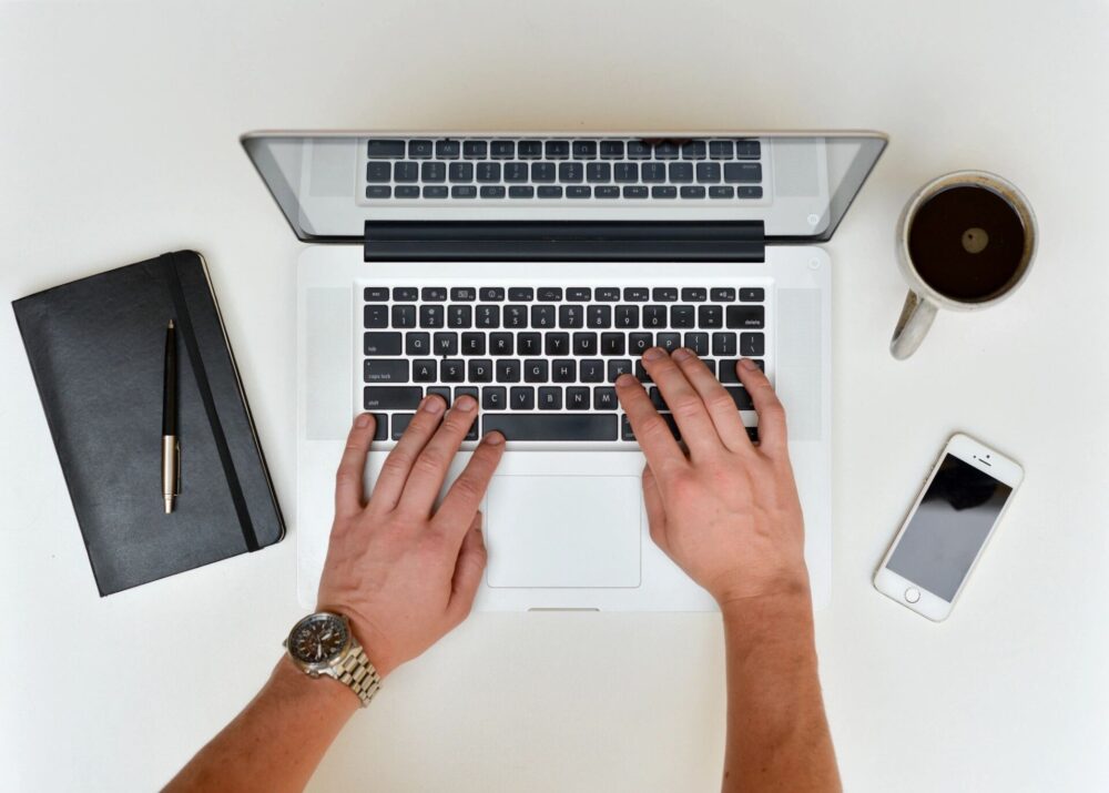 A person is working on a laptop with a notebook, pen, smartphone, and coffee nearby, suggesting a productivity or office setting.