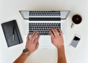 A person is working on a laptop with a notebook, pen, smartphone, and coffee nearby, suggesting a productivity or office setting.