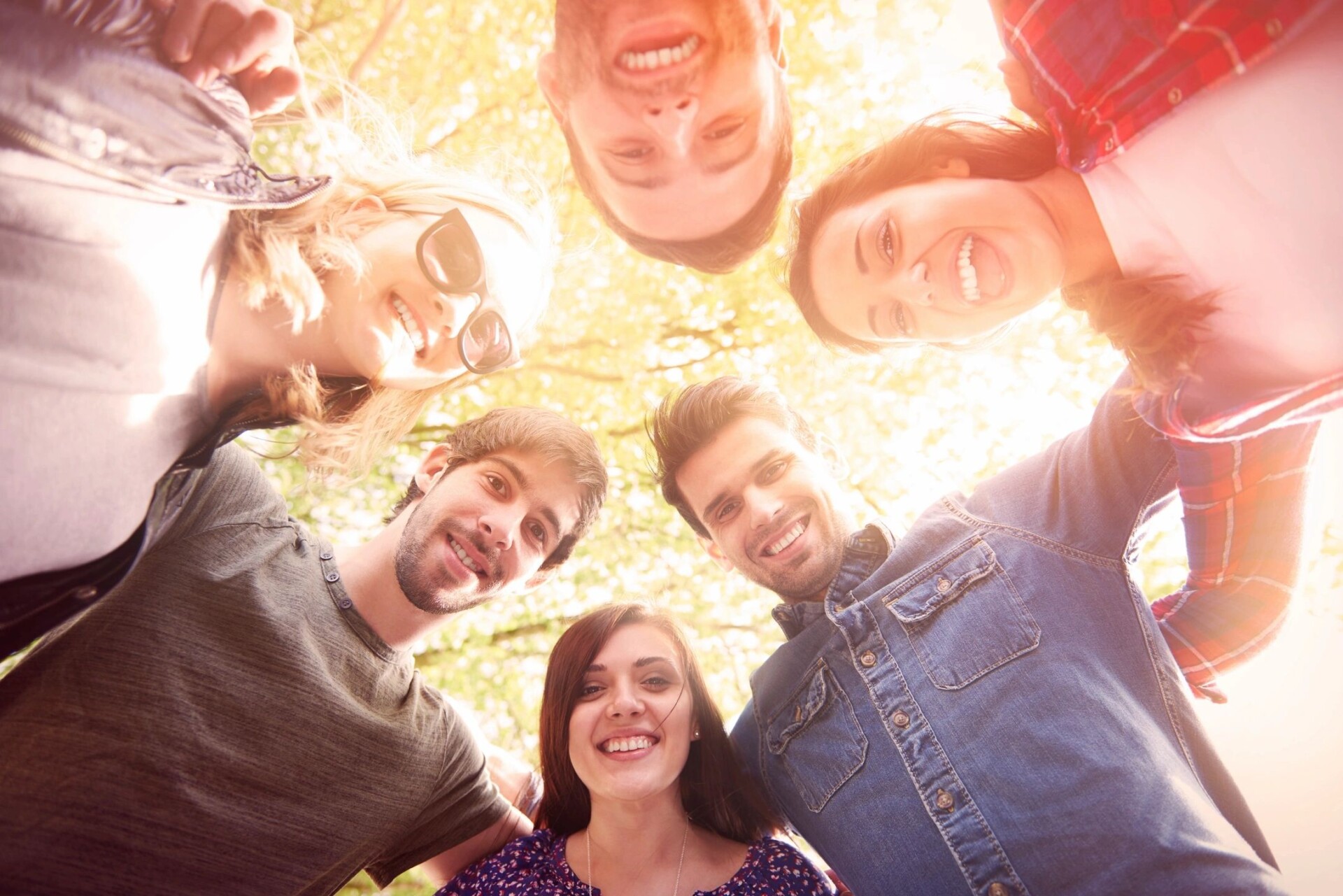 A group of five cheerful people huddle together, looking down at the camera, with sunlight and trees in the background.